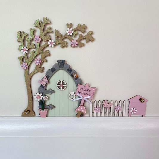 Beautiful Sage Green & Pink Fairy Door Set with Blossom Tree, Fence & Beehive  Invite some fairy magic into your home and capture your child’s imagination with this beautiful handmade wooden fairy door set.  All of our fairy door sets are designed and lovingly handcrafted. Each little door and accessory is hand painted and decorated with fairy glitter and embellishments.